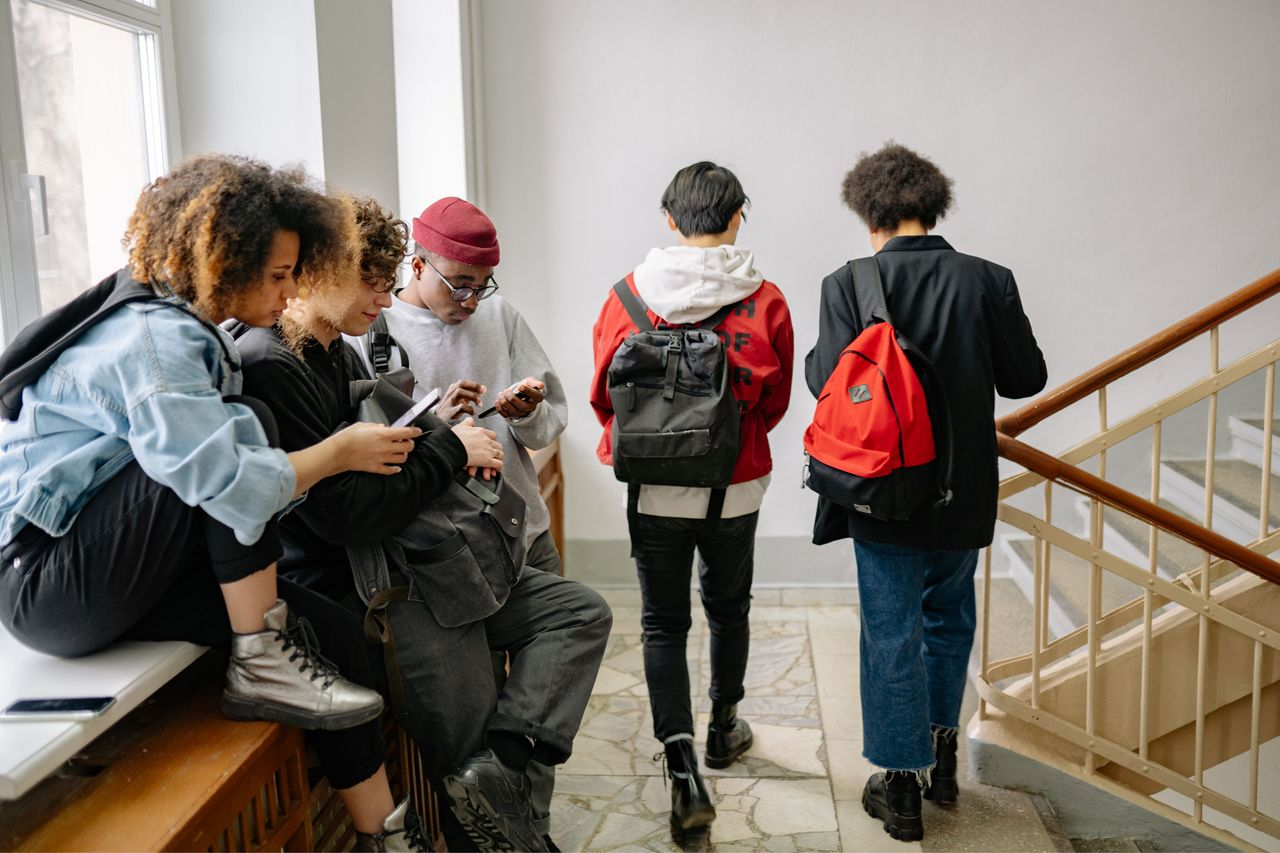A group of students sitting on close to a window on a staircase, using there phones. To the right of them, two students are walking by.